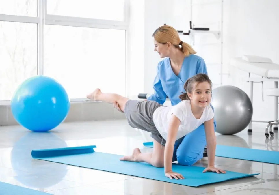 Physical therapist assisting young child in plank position on blue exercise mat in bright clinical space