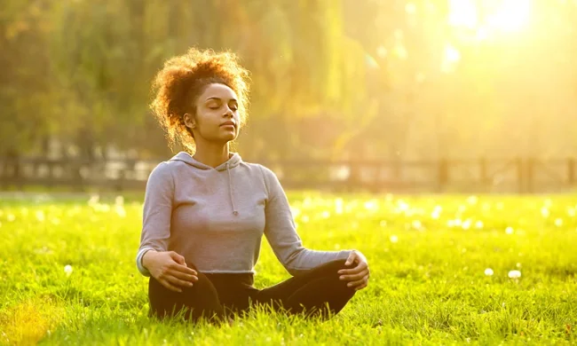 Woman with curly hair sitting cross-legged on grass in sunlit park, wearing gray long-sleeve shirt