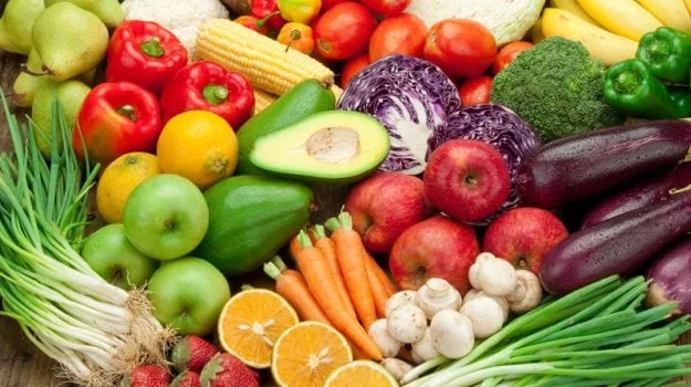 Assorted fresh vegetables and fruits including apples, peppers, broccoli, carrots, avocado, corn, tomatoes, eggplant, mushrooms, and citrus on wooden surface