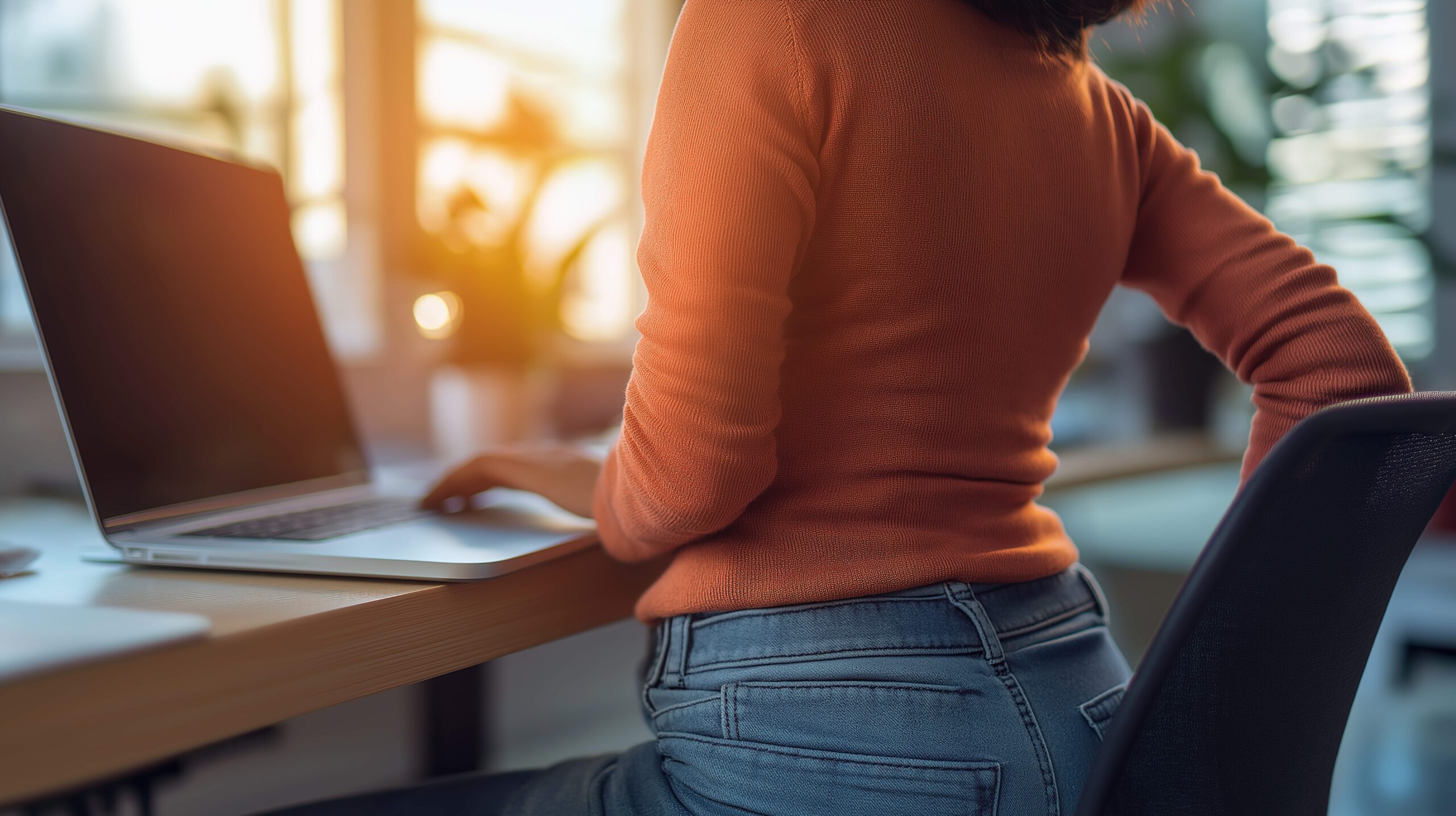 Summer Screen Time Posture Person wearing an orange sweater sitting at a desk with a laptop, experiencing back pain while working