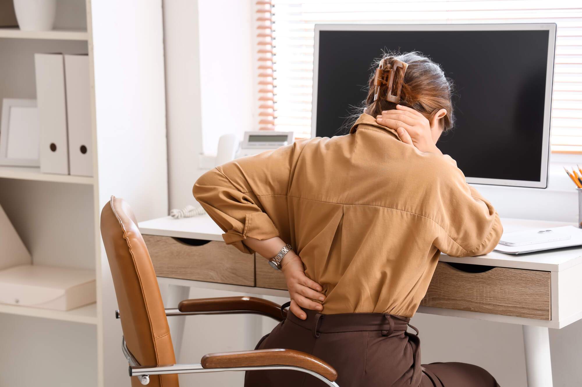Woman sitting at desk with hand on lower back, displaying signs of back pain while working at computer
