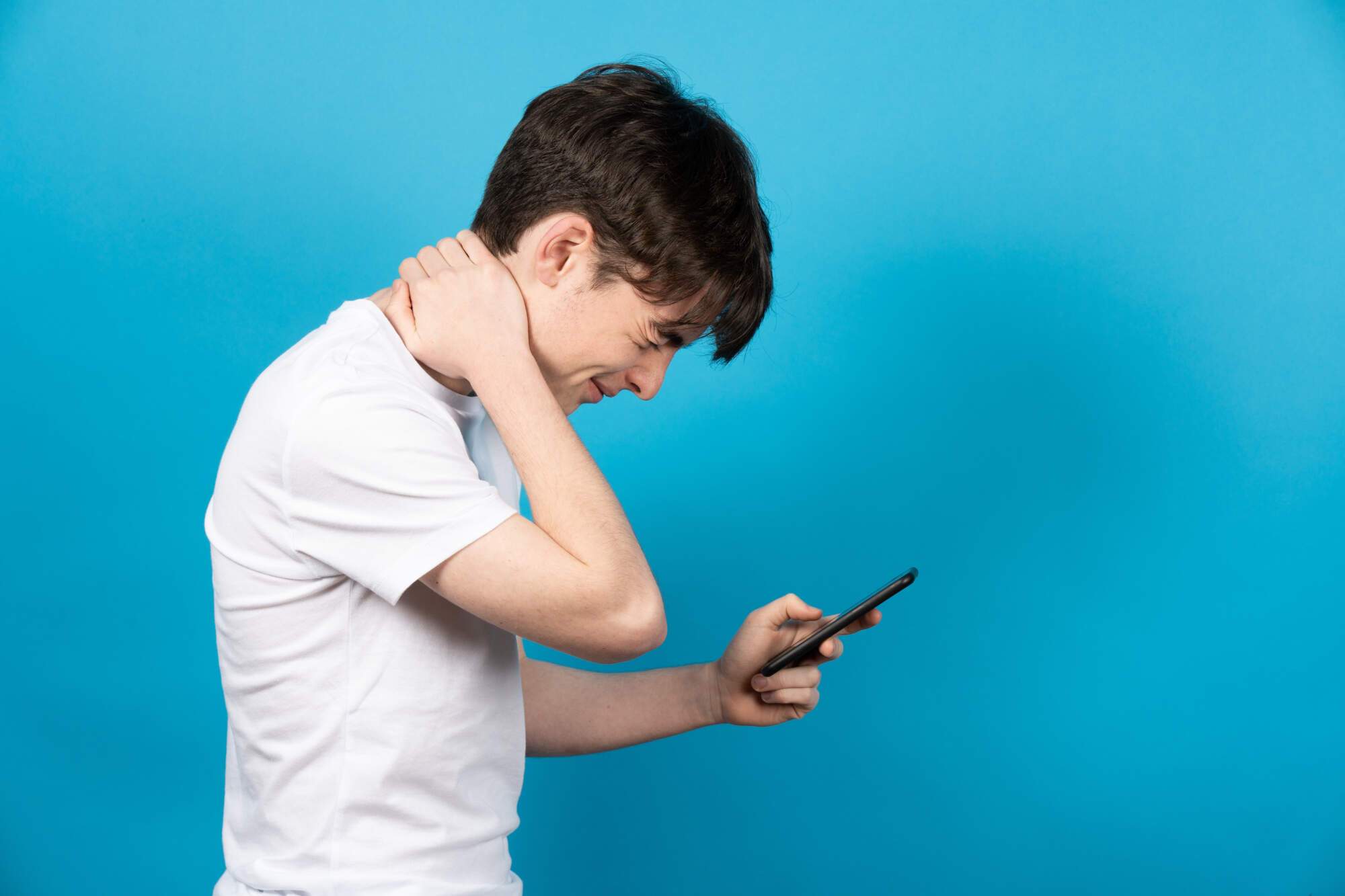 Teen Phone Posture Pain Teenage boy in white shirt holding smartphone at chest level, touching his neck in discomfort against a blue background.